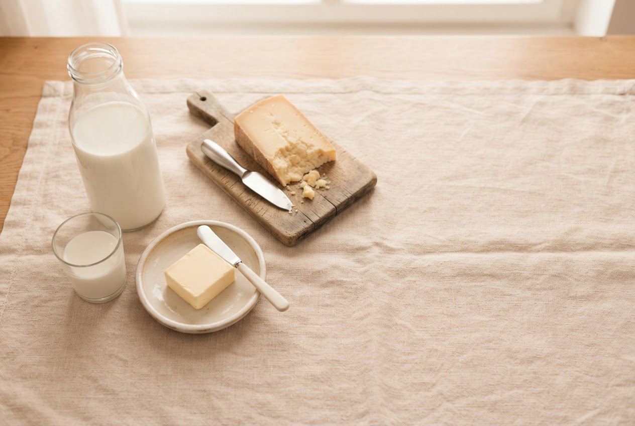 Eine Glasflasche Milch und ein gefülltes Glas, ein Stück Butter auf einem Teller und ein Keil Hartkäse auf einem Holzbrett, auf Leinen